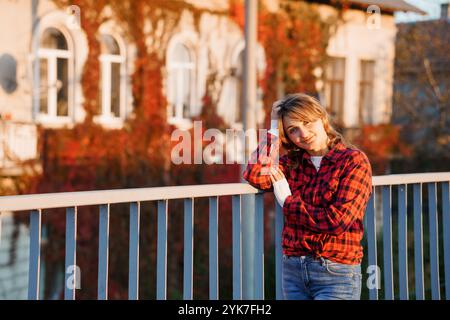 Jeune femme dans une chemise à carreaux rouge posant sur un balcon ensoleillé avec un bâtiment couvert de lierre en arrière-plan. Banque D'Images