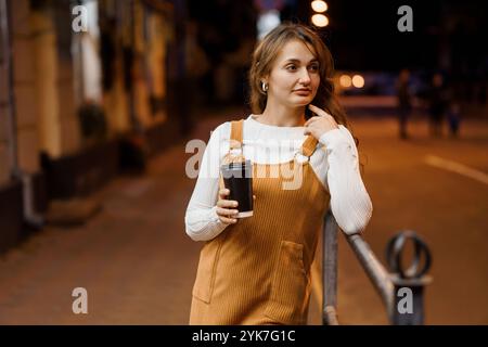 Jeune femme dans l'ensemble brun posant soigneusement avec tasse à café dans le cadre de nuit urbaine. Banque D'Images