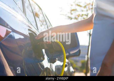 Close up femme main connecte un câble pour la charge à la voiture électrique contre la lumière du soleil. Banque D'Images