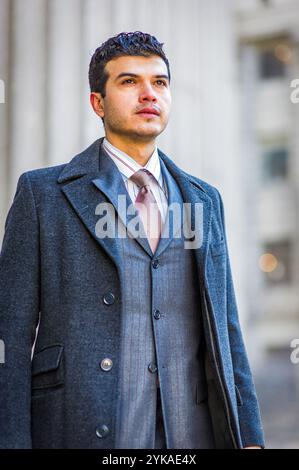 Un homme dans un manteau gris élégant et costume se tient en toute confiance à l'extérieur, regardant au loin. L'arrière-plan présente des éléments architecturaux flous t Banque D'Images