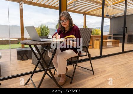 Femme d'affaires senior écrivant avec un stylo dans un cahier à côté d'un ordinateur portable sur une pergola de terrasse le matin à l'extérieur Banque D'Images
