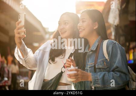 Deux jolies et joyeuses jeunes touristes asiatiques aiment explorer la nourriture de rue dans un marché en Thaïlande par une journée ensoleillée, prenant un selfie avec un sma Banque D'Images