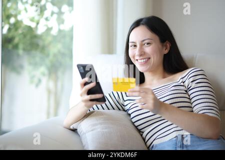 Heureuse femme asiatique achetant en ligne tenant le téléphone et la carte de crédit regardant la caméra assis sur un canapé à la maison Banque D'Images