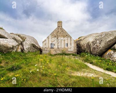 Une petite maison en pierre nichée entre deux grands rochers à Kerlouan, dans un paysage herbeux sous un ciel nuageux. Banque D'Images