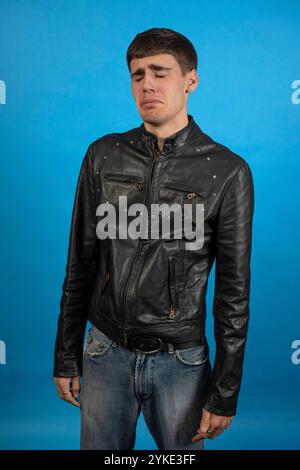 Triste jeune homme pleurant à cause de problèmes, portant une veste en cuir noir et un Jean sur un fond bleu Banque D'Images