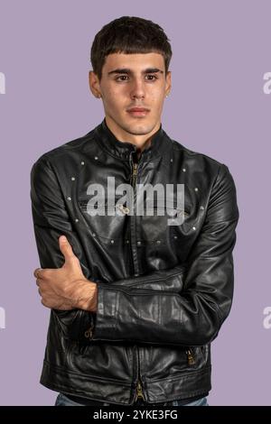 Photo studio d'un jeune homme à la mode portant une veste en cuir noir posant avec les bras pliés sur fond violet Banque D'Images