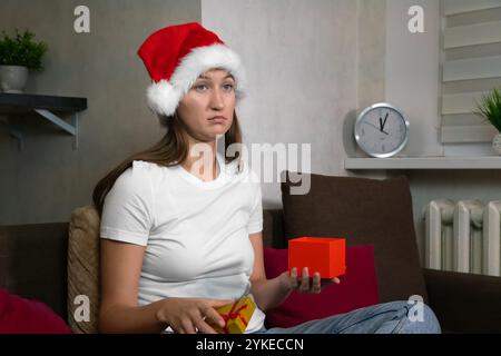 Femme triste portant chapeau de père noël ouvrant la boîte cadeau de noël à la maison, exprimant sa déception avec le cadeau qu'elle a reçu Banque D'Images