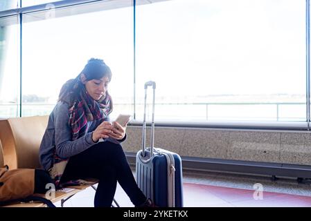 Femme assise dans la salle d'attente de l'aéroport tout en utilisant un téléphone portable Banque D'Images