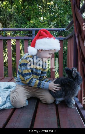 Garçon mignon et lapin portant chapeau de Père Noël sur la terrasse Banque D'Images