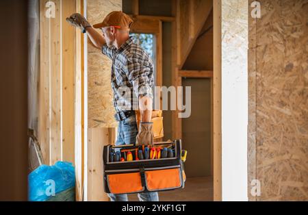 Un ouvrier de la construction se tient à côté d'un mur de bois, tenant une boîte à outils remplie de divers outils, se préparant à travailler dans une maison en construction. Banque D'Images