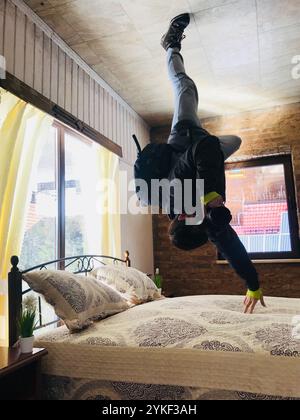 Un homme exécute un mouvement de breakdance dynamique sur le plafond d'une chambre élégante, créant une illusion visuelle intrigante. La chambre est lumineuse et bien déco Banque D'Images