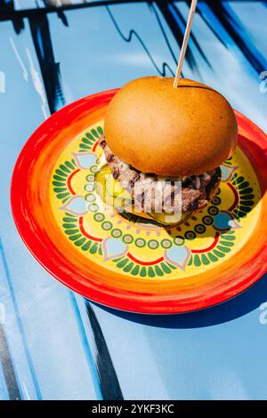 Un succulent burger de bœuf avec des cornichons et du fromage, présenté sur une assiette en céramique colorée, posé sur une surface de table bleue qui ajoute une touche moderne à t Banque D'Images