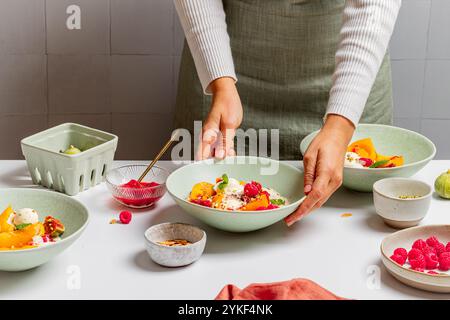 Une personne garnissant habilement un bol de dessert pêche garni de crème glacée, entouré de bols de différentes garnitures et de fruits frais sur un compte de cuisine Banque D'Images