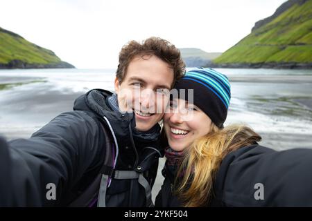 Couple vêtu d'un équipement de plein air chaud prenant un selfie à la plage de sable noir de Tjørnuvik dans les îles Féroé les collines verdoyantes et dramatique la côte Banque D'Images
