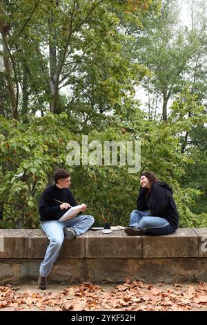 Un couple LGBTQIA+ partage un moment serein à l'extérieur, entouré d'arbres luxuriants et l'automne laisse Un homme trans esquisser tout en s'engageant dans la conversation, em Banque D'Images