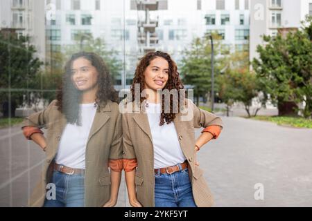 Une femme d'affaires brésilienne joyeuse dans une tenue élégante se tient en toute confiance à l'extérieur d'un bâtiment moderne, reflétant positivité et professionnalisme Banque D'Images