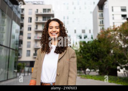 Une femme d'affaires brésilienne joyeuse aux cheveux bouclés, portant un blazer à carreaux élégant, se tient confiante dans un fond urbain moderne, représentant des profes Banque D'Images