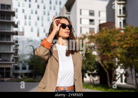 Femme d'affaires brésilienne confiante dans une tenue décontractée d'affaires à la mode, ajustant les lunettes de soleil, avec des bâtiments modernes en arrière-plan Banque D'Images