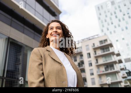 Une femme d'affaires brésilienne professionnelle aux cheveux bouclés, vêtue d'un costume élégant, sourit en toute confiance tout en se tenant à l'extérieur avec des bâtiments modernes dedans Banque D'Images