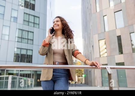 Femme d'affaires brésilienne souriante dans une tenue de bureau décontractée s'engageant dans une conversation joyeuse sur son téléphone portable tout en se tenant à l'extérieur dans une ville moderne Banque D'Images