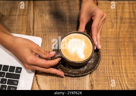Une image en gros plan capturant les mains d'une femme d'affaires tenant un latte crémeux à côté de son ordinateur portable sur une table en bois rustique Banque D'Images