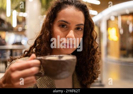 Une femme d'affaires brésilienne confiante sirote un café dans un café animé, mettant en valeur son attitude tranquille dans un cadre décontracté Banque D'Images