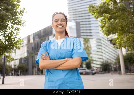Une jeune étudiante infirmière se tient confiante, les bras croisés dans un environnement urbain, portant un gommage bleu et souriant chaleureusement Banque D'Images