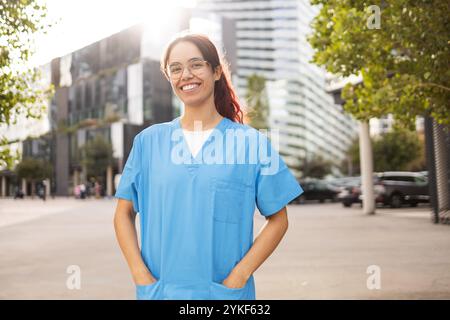 Une jeune femme joyeuse dans des gommages d'infirmière bleus se tient en toute confiance à l'extérieur, incarnant l'optimisme et le professionnalisme dans un cadre urbain moderne Banque D'Images