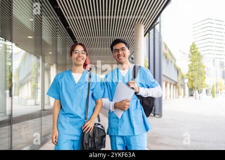 Un jeune homme et une jeune femme vêtus d'uniformes d'infirmière bleus, souriant et tenant des papiers, marchent en toute confiance à l'extérieur d'un bâtiment universitaire moderne Banque D'Images