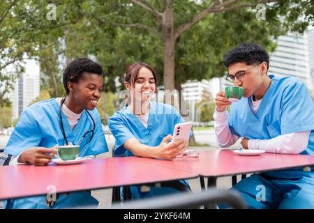 Un groupe de trois étudiants en soins infirmiers, vêtus de gommages bleus, profitent d'une pause à l'extérieur on rit et tient un smartphone, un autre boit du café, tandis que le Banque D'Images
