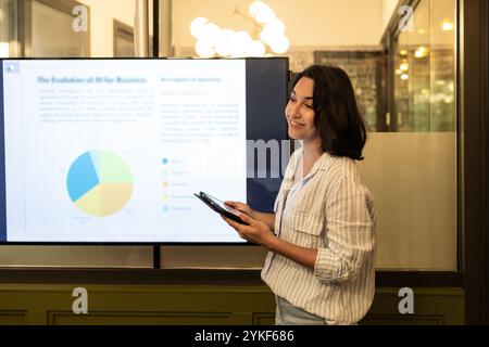Une femme dans un espace de coworking moderne présente des données sur un écran elle tient une tablette, expliquant avec confiance ses points de vue, mettant en évidence la collaboration et la cr Banque D'Images