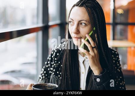 Une femme caucasienne élégante avec des tresses parle sur son téléphone portable tout en sirotant un café dans un café moderne. Elle regarde loin de la caméra, exsudant une détente Banque D'Images
