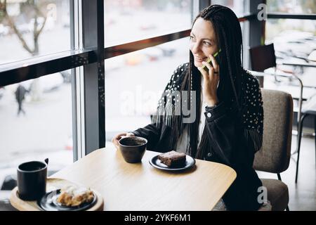Une femme caucasienne élégante avec des tresses sourit tout en parlant au téléphone et en buvant du café dans un café. Elle porte une veste cloutée et est assise par un W. Banque D'Images