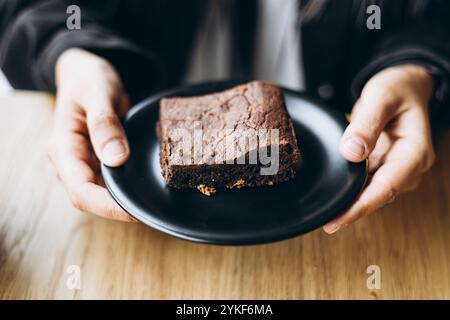 Une main recadrée méconnaissable d'une personne alors qu'ils tiennent une assiette avec un brownie au chocolat fraîchement cuit. L'accent est mis sur la partie supérieure texturée du Banque D'Images