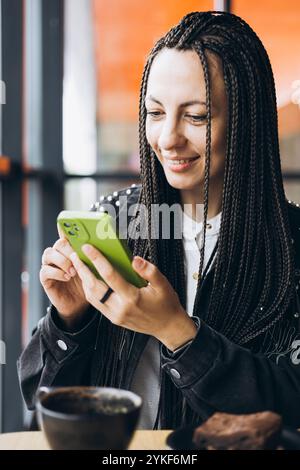 Une femme caucasienne élégante avec des tresses sourit tout en utilisant un smartphone et en buvant du café dans un café. Elle porte une veste noire et est fiancée avec Banque D'Images