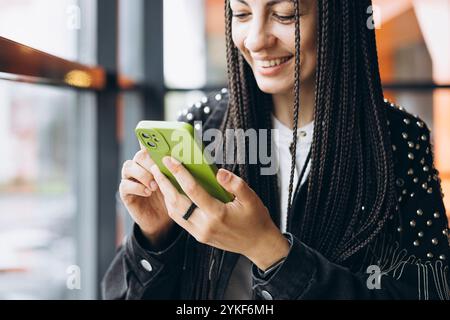 Une femme caucasienne élégante avec des tresses sourit alors qu'elle utilise son smartphone dans un café, affichant une scène de style de vie décontracté et moderne. Banque D'Images