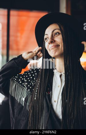 Une femme caucasienne élégante aux cheveux tressés sourit doucement alors qu'elle ajuste son chapeau noir, debout dans un cadre chaleureusement éclairé avec un sourire subtil. Banque D'Images