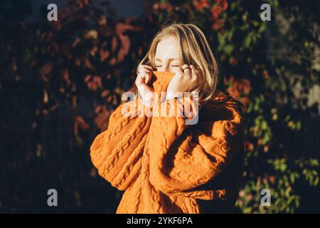 Une femme se prélasse dans la chaleur d'un pull orange tricoté confortable, englouti dans la lumière du soleil avec un fond de feuillage automnal, incarnant le confort et la esse Banque D'Images