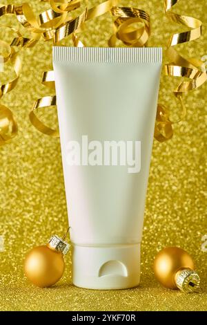 Un tube cosmétique placé au milieu de confettis dorés étincelants et d'ornements de Noël sur un fond doré chatoyant. Le tube est sans marque, ce qui le rend ide Banque D'Images