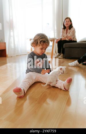 Dans une pièce chaleureusement éclairée, un jeune enfant est assis sur le plancher en bois, caressant soigneusement un petit chaton blanc, avec une femme adulte souriante observant d'un di Banque D'Images