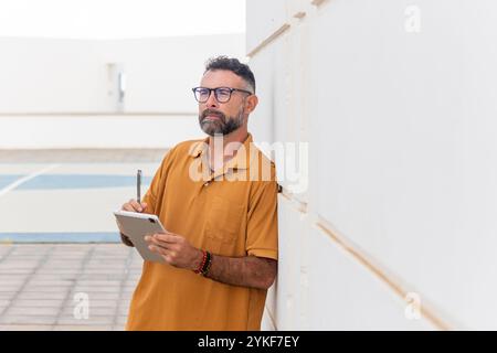 Un homme gay pensif d'âge moyen avec une barbe, des lunettes, et portant une chemise décontractée se tient près d'un mur extérieur lumineux, tenant une tablette et un stylo numérique, Banque D'Images