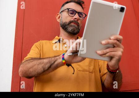 Un homme gay d'âge moyen avec une barbe, portant des lunettes et un bracelet arc-en-ciel, axé sur l'utilisation d'une tablette sur un fond rouge Banque D'Images