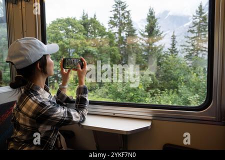 Une touriste asiatique capture les paysages luxuriants de Lauterbrunnen, en Suisse, en utilisant son smartphone dans le confort d'un train en mouvement sur lequel elle se concentre Banque D'Images