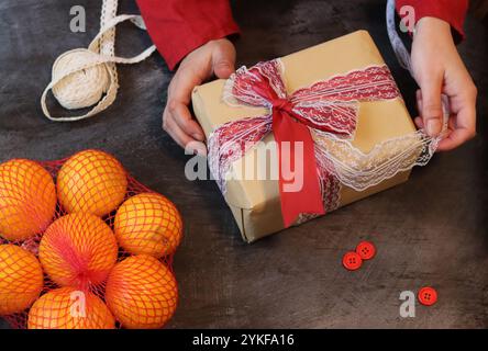 Mains d'une jeune femme portant une robe rouge enveloppant des cadeaux de Noël sur un fond en bois. Boîte cadeau avec ruban rouge et mandarines vue du dessus photo Banque D'Images