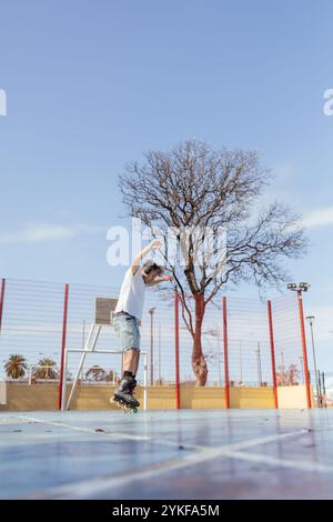 De dessous, un jeune homme avec des écouteurs est capturé à mi-saut tout en faisant du roller avec des patins à roues alignées sur un terrain de sport extérieur. Concentré sur son patinage, w Banque D'Images