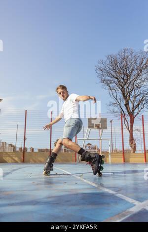 d'en bas, un jeune homme est capturé en action alors qu'il roule sur des patins à roues alignées sur un terrain de sport ensoleillé, se concentrant intensément sur ses mouvements. Banque D'Images