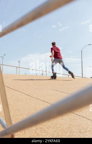 Vue arrière d'un jeune homme méconnaissable en roller sur des patins à roues alignées, capturé sur un pont sous un ciel bleu vif. Banque D'Images