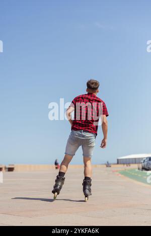 Vue arrière d'un jeune homme méconnaissable roller par une journée ensoleillée, vêtu d'une chemise à carreaux rouges et d'un short en Jean. L'image montre l'énergie A. Banque D'Images