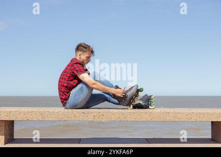 Un jeune homme concentré est assis sur une barrière de béton près de la plage, ajustant soigneusement ses patins à roues alignées. Il est habillé d'une chemise à carreaux rouge et noir A. Banque D'Images