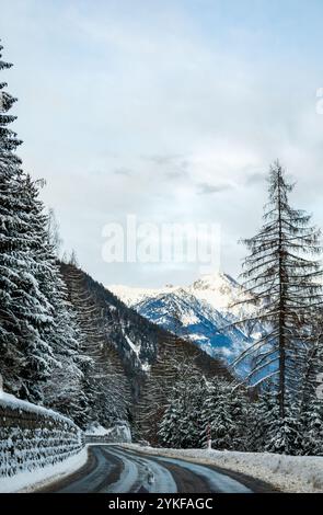 Une route sinueuse serpente à travers les forêts enneigées des Alpes, encadrées par des montagnes majestueuses sous un ciel hivernal serein, capturant l'essence de Banque D'Images